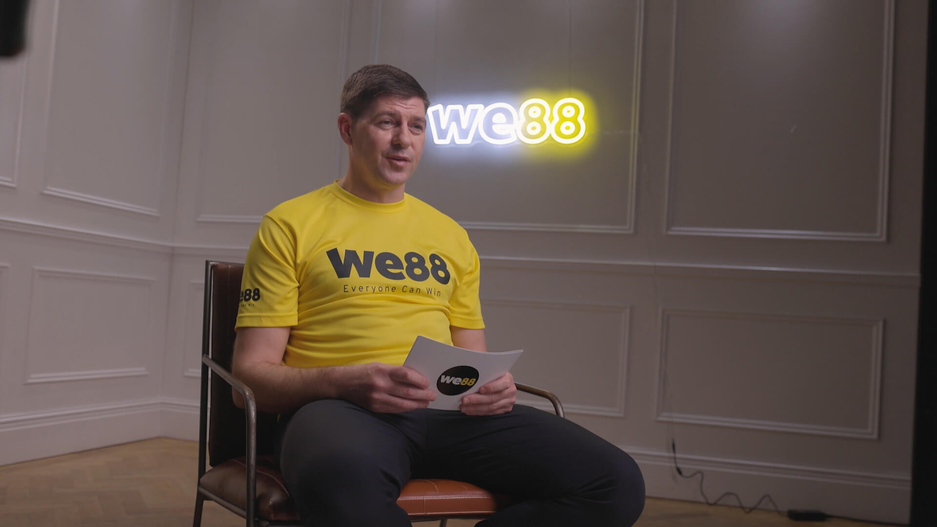 Steven Gerrard sitting in a leather chair wearing a yellow WE88 official jersey, talking to the camera and holding a WE88 script