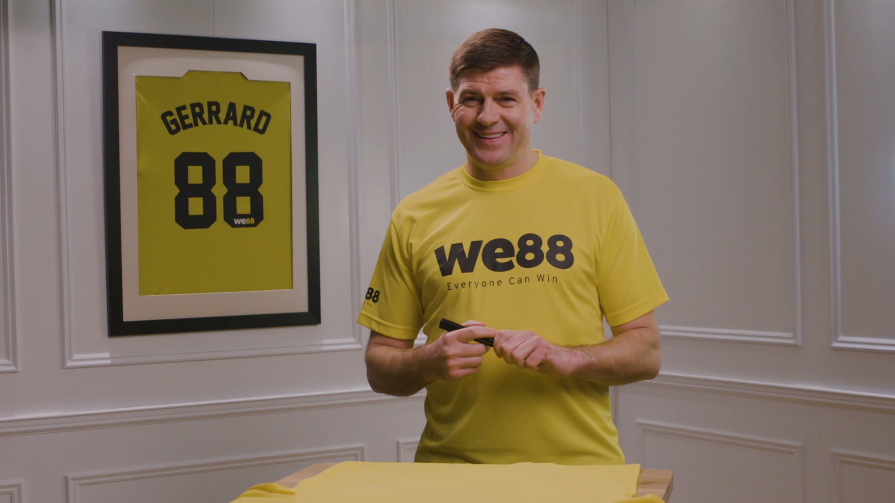 WE88 26/27 Global Brand Ambassador Steven Gerrard smiling with a marker, preparing to sign a jersey during a behind-the-scenes shoot