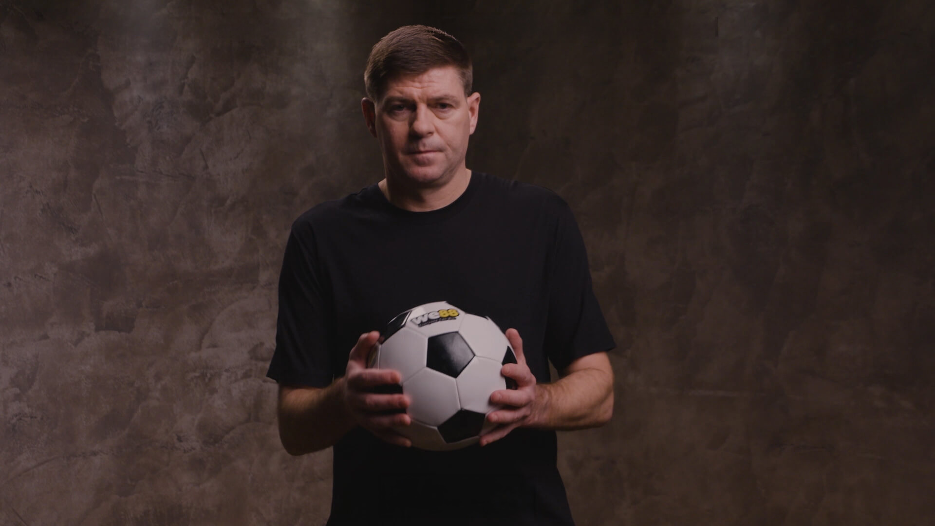 WE88 26/27 Global Brand Ambassador Steven Gerrard holding a custom WE88 football during a dramatic studio photoshoot